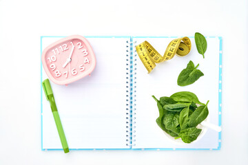 Composition with  spinach, measuring tape, blank paper notebook and pink alarm clock on white background. Mock up. Diet concept, copy space