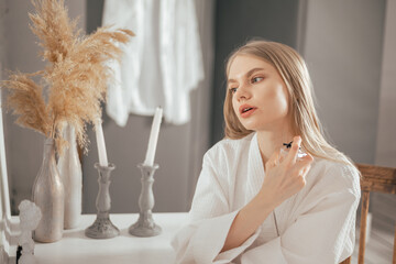Young blonde beautiful woman in the white bathrobe sitting next to the table and using perfume...
