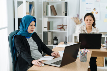 Plakat Young business people working together at office. muslim pregnant woman in headscarf sitting at workplace typing on laptop computer with colleagues concentrated using notebook pc in background.