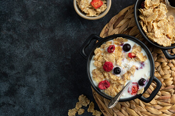 Corn flakes with strawberry and blueberry in black bowls over dark background, copy space
