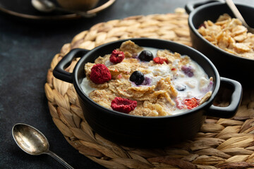 Corn flakes with strawberry and blueberry in black bowls over dark background, top view.