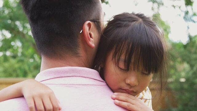 Loving Asian father cuddling tired daughter in garden as girl looks over his shoulder - shot in slow motion