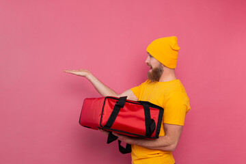 European bearded delivery man with box with food smiling and pointing finger to left on pink background isolated