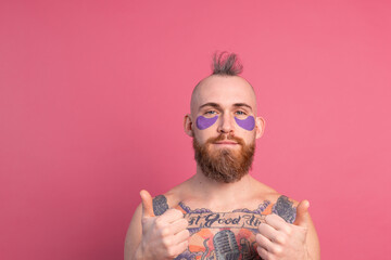 European handsome bearded tattooed topless man with purple eye patches mask posing to camera, face care for men. Isolated on pink background.