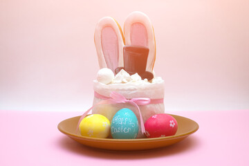 White cake with bunny ears and black hat. in brown plate with three colorful eggs on the white and pink background