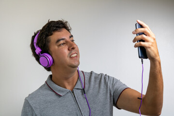 Brunette man listening to music at home with purple headphones and a cellphone. Wearing a gray polo shirt on a white background. Happiness and authenticity in a relaxing time.