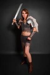 Medieval woman warrior in chain mail armor with lamellar bracers and lamellar shoulder pads with polar fox fur on her shoulders stands with a sword in her hands against a dark background.