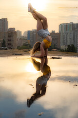 Flexible female gymnast doing handstand and calisthenic with reflection in the water on cityscape background during dramatic sunset