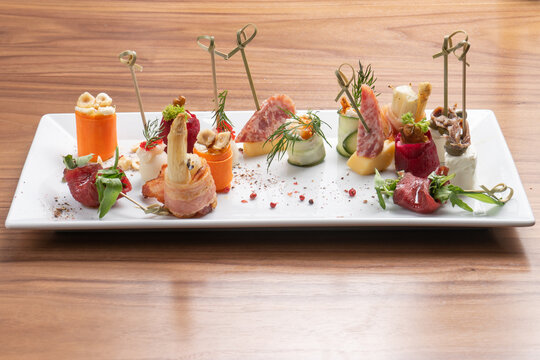 An assortment of many small meat snacks to accompany the wine. Canapes on skewers of zucchini, carrots, bacon, sausage and cheese in a white rectangular ceramic plate on a wooden table.