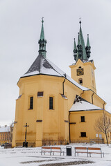 Yellow baroque church of St. James the Elder with gothic clock tower, medieval historical buildings, Main town square under snow in winter day, Pribram, Bohemia, Czech Republic
