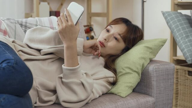 lazy ambience korean girl reclining on sofa phubbing smartphone feels bored and yawns. spiritless asian woman doing nothing but using her phone in living room didn't see anything interesting.