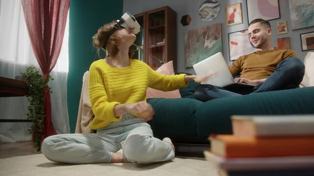 Young happy woman having fun playing VR headset video games and use technology virtual reality at home, man with laptop share ar content to girlfriend