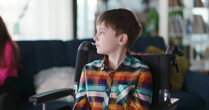 Portrait of unwell caucasian disabled child sitting in wheelchair looking at camera. His happy old er sister having fun playing with socilal media on smartphone. Diversity.
