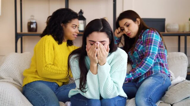 Sad Arabic woman sitting on sofa and closing face with hands, multiethnic roommates gossiping behind her back. Evil females lacking sympathy for friend in stress. Concept of emotion