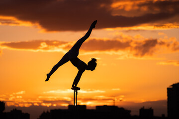 Silhouette of female circus artist doing handstand on the dramatic sky background during sunset