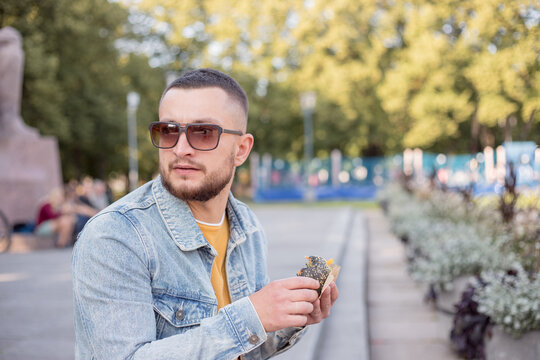 Hipster Man Eating Burger On The Street. Young Bearded Stylish Guy Biting Delicious Burger.Close Up Of Young Man Eating Burger Outdoors. Man With Beard Having Street Food.