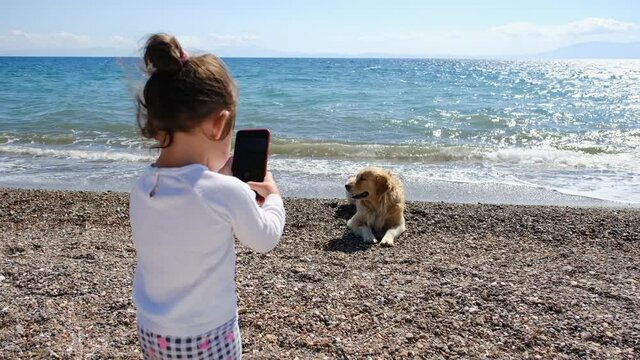 Little girl taking pictures of her dog on the beach with her mobile phone. Generation Z