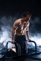 Muscular build man doing calisthenics on parallels bar indoor on black, smoked background. concept...