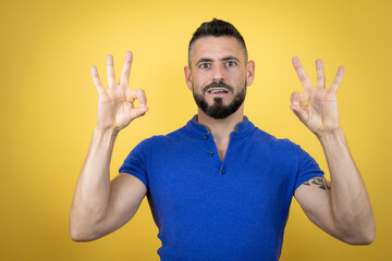 Handsome man with beard wearing blue polo shirt over yellow background doing ok sign with fingers and smiling, excellent symbol