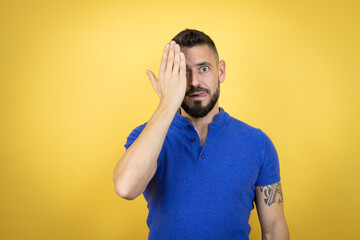 Handsome man with beard wearing blue polo shirt over yellow background covering one eye with hand, confident smile on face and surprise emotion.