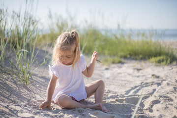 Little girl at the beach, summer dressed child girl sit down on sand beach playing smiling with sand, warm colours
