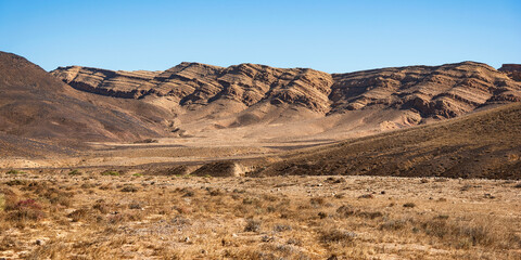 tilted limestone cliff of the south rim of the Makhtesh Ramon crater in Israel between two ancient volcanic hills against a clear sky