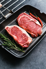 Raw rib eye and striploin New York beef steak on black grilling pan with chili pepper and rosemary, top view.