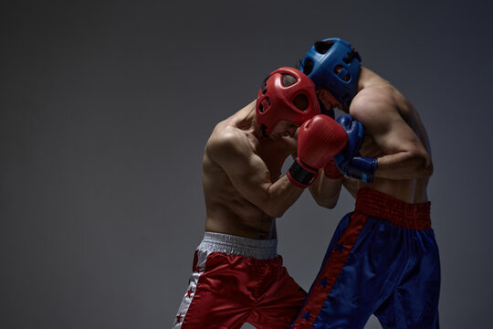 Sparring of two fighting males in helmets and boxing gloves on gray studio background, martial arts, mixed fight concept