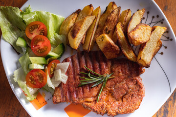 Grilled pork with baked potatoes and with salad. The food is served on a plate. Delicious grilled meat. Roasted pork steak on the white plate on the wooden background.