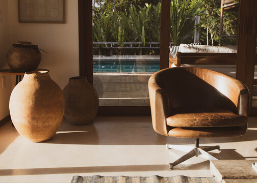 Modern luxury summer holiday or vacation beach house living room interior with rustic ceramic clay vases and armchair on the foreground and view of the swimming pool through the glass door.
