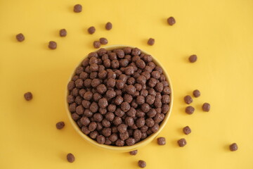 Chocolate cereal corn balls in a yellow bowl scattered on a yellow background. Top view. Copy, empty space for text