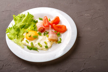 Scrambled eggs with bacon garnished with tomato and green onion on a gray concrete background.