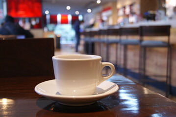 Tasa de café blanca sobre mesa de madera con restaurante bar al fondo (foco en tasa)