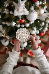 Woman with cup of delicious hot drink near Christmas tree at home, top view