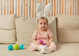 baby girl in a dress and rabbit ears sits on a sofa with colorful eggs