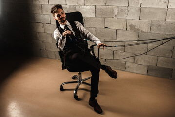 frightened, worried, pensive, man sitting on a chair, testing on a polygraph, wearing polygraph sensors, sitting on a gray brick wall background, wires held in his hands, holding hands, top view.