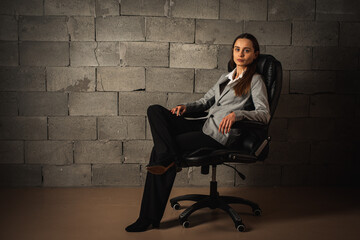 beautiful, stylish, confident businessman lady, brunette in a suit sits on a leather chair and looks straight, on a gray background. low key. Selective focus