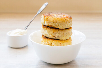 Homemade cottage cheese pancakes syrniki stack on round glossy white bowl, sauce pan, sour cream, spoon on white rustic natural wooden table background. Close up. Selective focus. Copy space