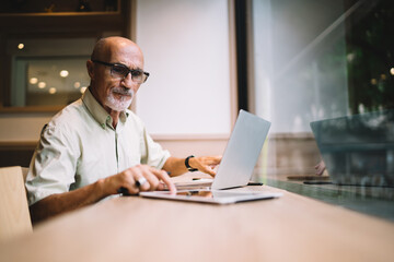 Aged freelancer in classic eyeglasses browsing received information on digital tablet sitting at desk with laptop computer, mature Caucaisan man 60s reading news and notifications on touch pad