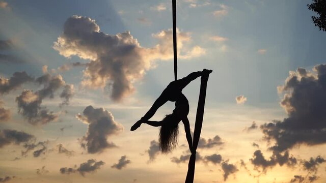 Female circus artist showing her flexibility and splits with red aerial silk on the sky background. Concept of healthy lifestyle