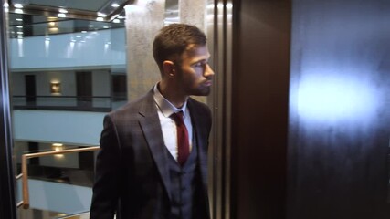 Close-up of handsome man wearing chic three-piece suit and tie getting out of transparent elevator car and walking along hotel corridor. Respectable male with travel bag taking lift down