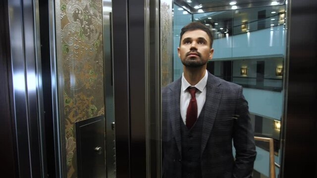 Close-up of handsome bearded businessman with travel bag using hotel guest's card while taking transparent lift up. Confident formally dressed male going elevator car up to check into hotel room