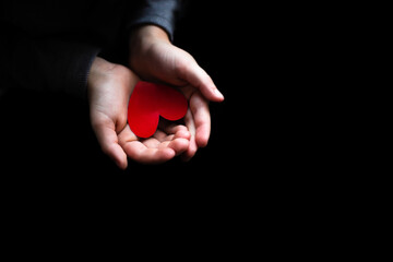 The child holds a red heart in his palms, hands close-up, black background, low key. Valentine's...