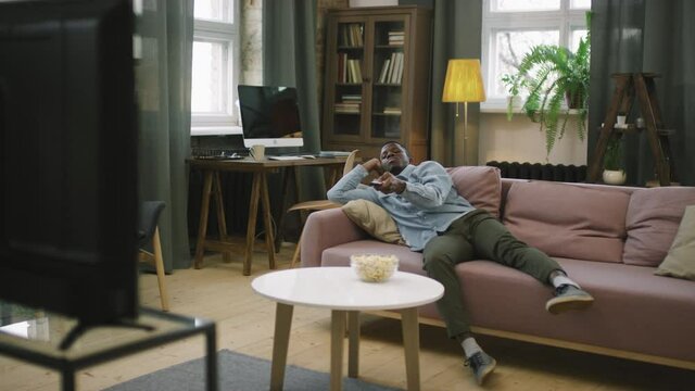 Full slow-motion arc shot of bored young afro man lying on couch in modern apartment switching channels on TV procrastinating