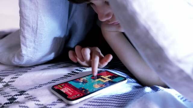 kid, a boy of 8 years old lies in bed under the covers, plays in the application on a smartphone, the concept of addiction, insomnia