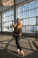 Fototapeta premium A stylish girl in a brown sweater, black pants with a large women's bag poses against the background of large windows.