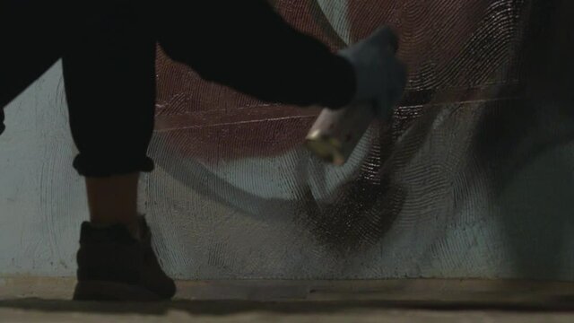 Artist in black hoodie, black shoes and blue gloves draws on the blue wall with spray paint inside building