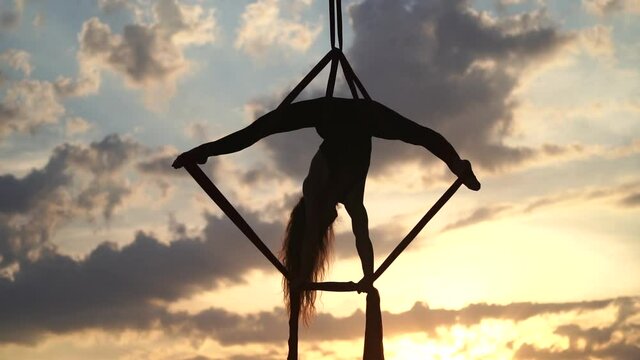 Silhouette of Female circus artist showing her flexibility and splits with red aerial silk on the dramatic sky background. Concept of movement and motion 