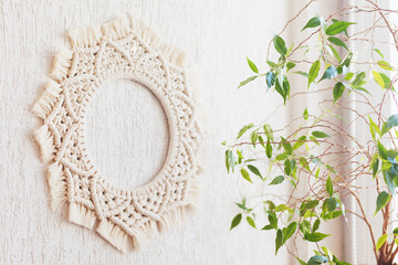 Cotton macrame mandala wall decoration hanging on white wall with green leaves. Handmade macrame wreath. Natural cotton thread. Eco home decor.  Copy space