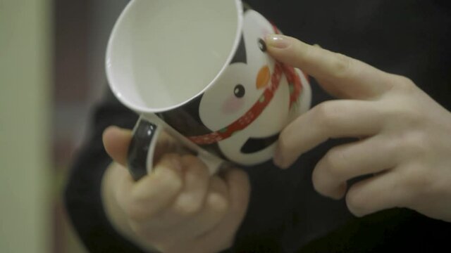 Woman in black blouse with clean nails holds an empty cup with a picture of a penguin. She looks at it from all angles, turning it in her hands.Closeup